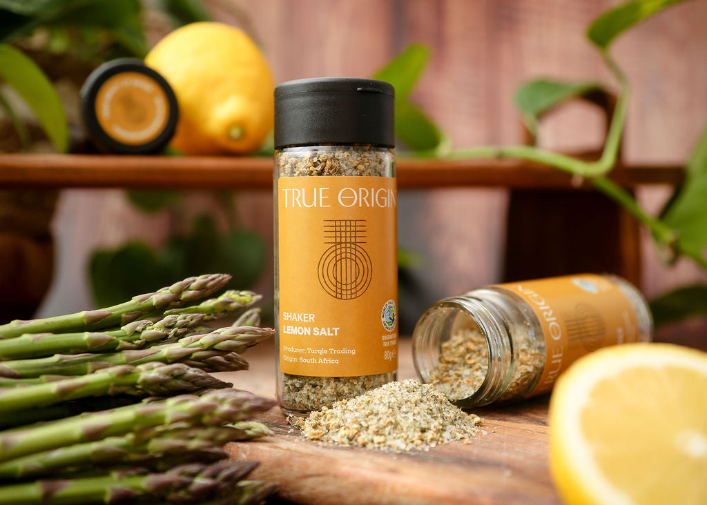 A yellow glass jar of lemon salt with a black lid and an open jar on its side with lemon salt pouring out onto a chopping board next to some asparagus and a lemon in the background.