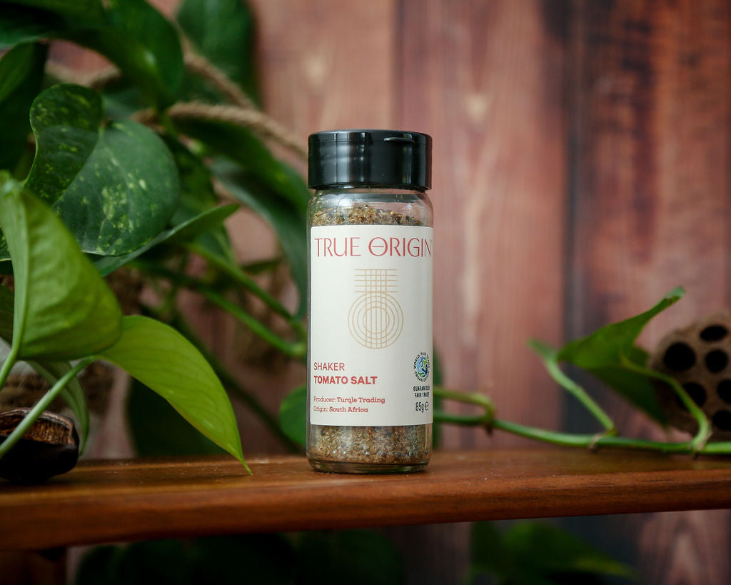 A white glass jar of tomato salt with a black lid on a wooden shelf with some green leaves in the background.