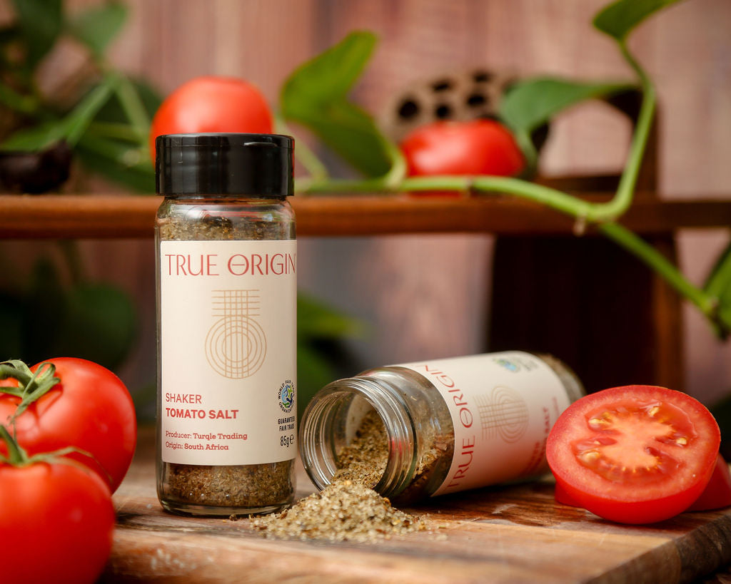 A white glass jar of tomato salt with a black lid beside a jar of fair trade tomato salt on its side with tomato salt pouring onto a chopping board next to some tomatoes.