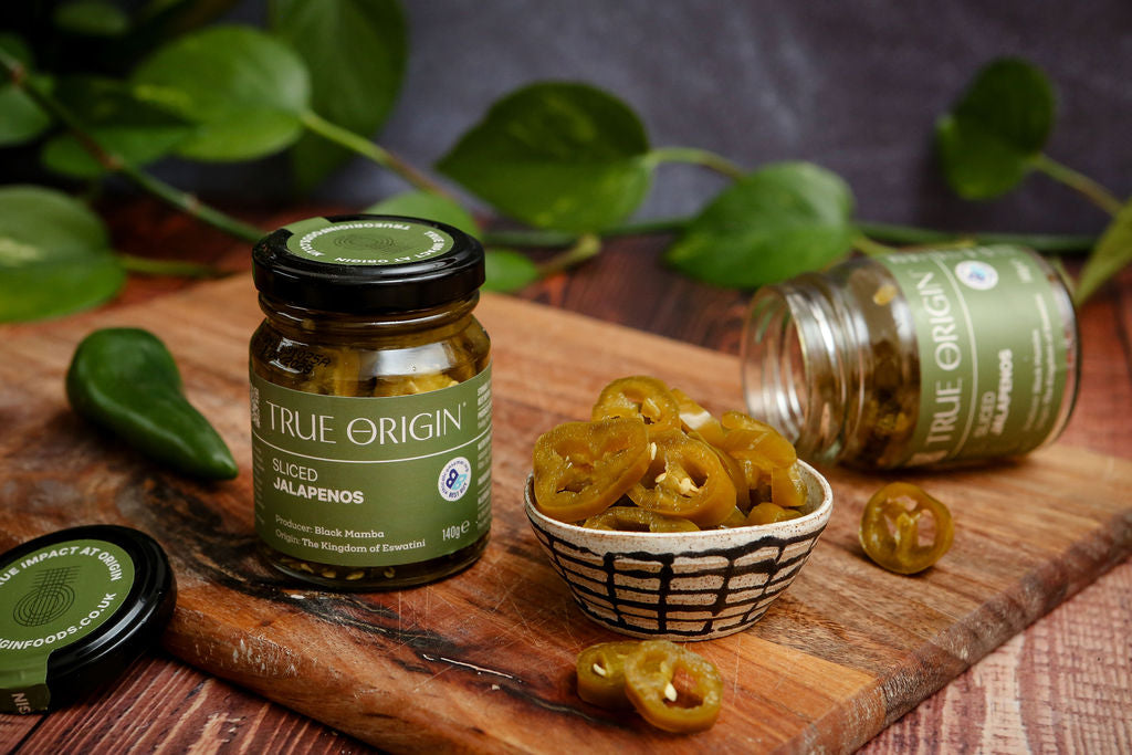 Jars of True Origin pickled jalapeños on a wooden board, along with a bowl of contents, with green leaves in the background.