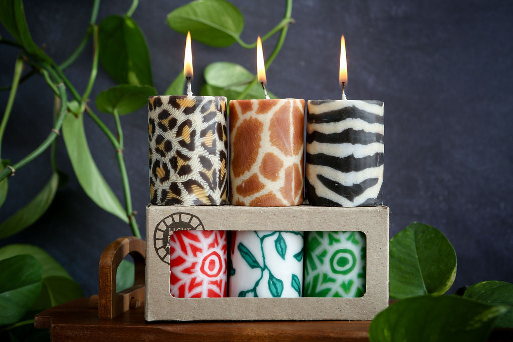 Set of 3 animal and 3 festive patterned candles in a cardboard holder with green leaves in the background