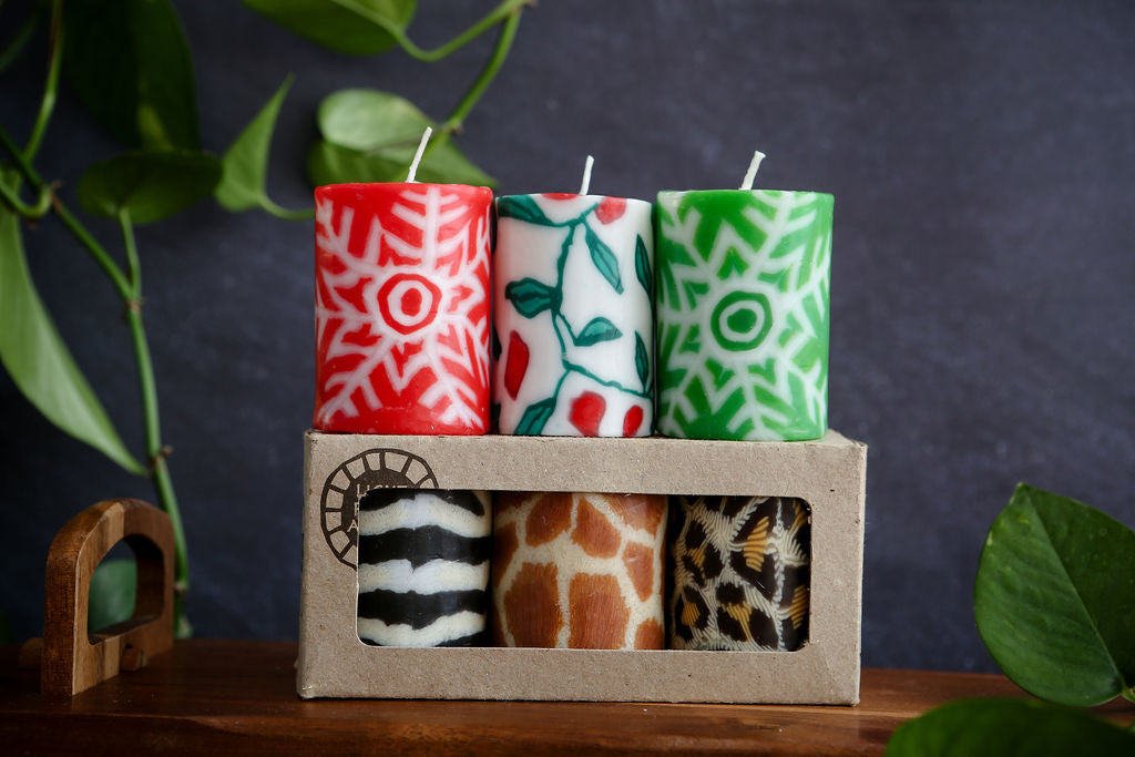 Set of 2 festive and 3 animal patterned candles in a cardboard holder with a dark background and green leaves.