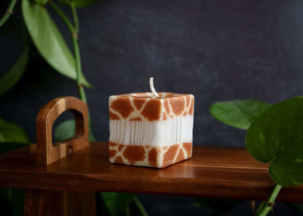 Square candle with giraffe pattern on a wooden surface with green leaves in the background