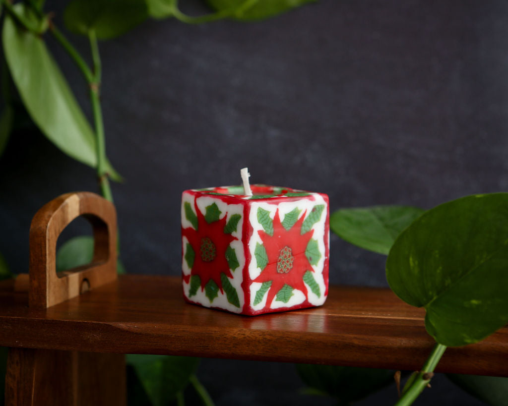 Decorative square candle with red and green pattern on a wooden surface with plants in the background
