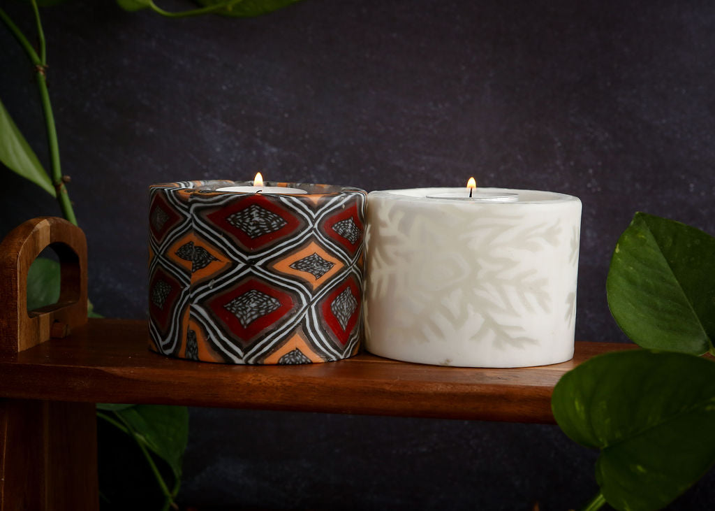 Two tealight holder candles, one with an African pattern and one white with a large snowflake, on a wooden surface with a dark background.