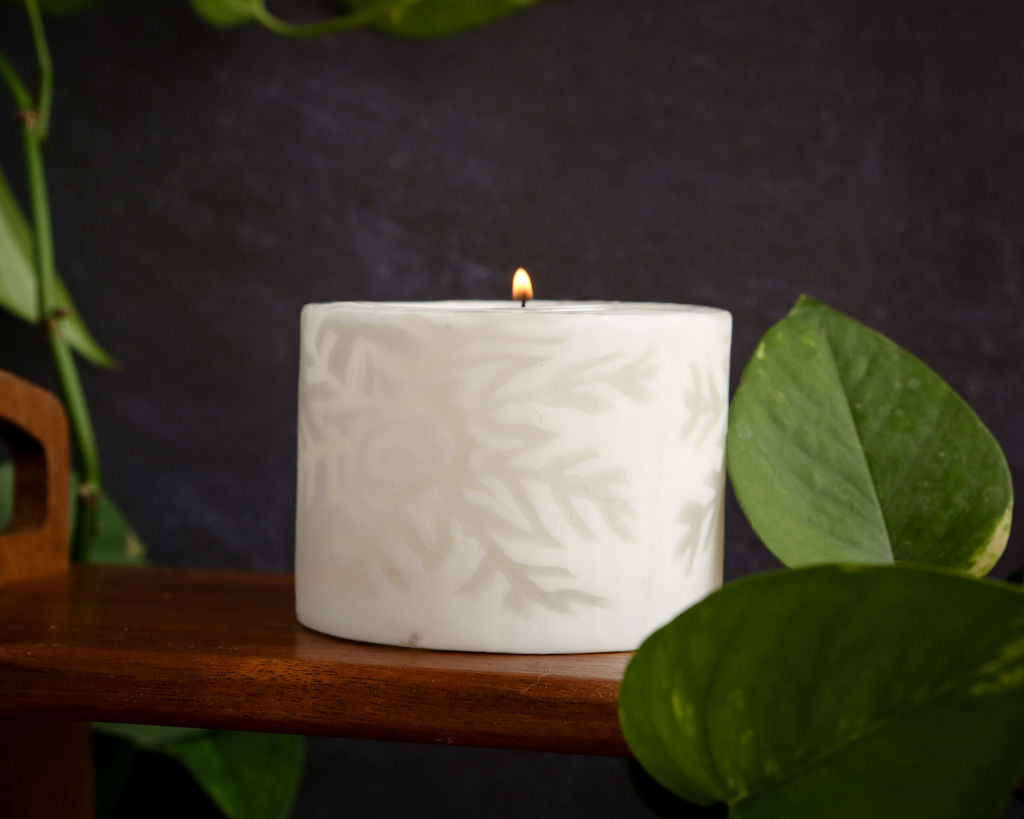 White textured candle on a wooden surface with green leaves in the foreground