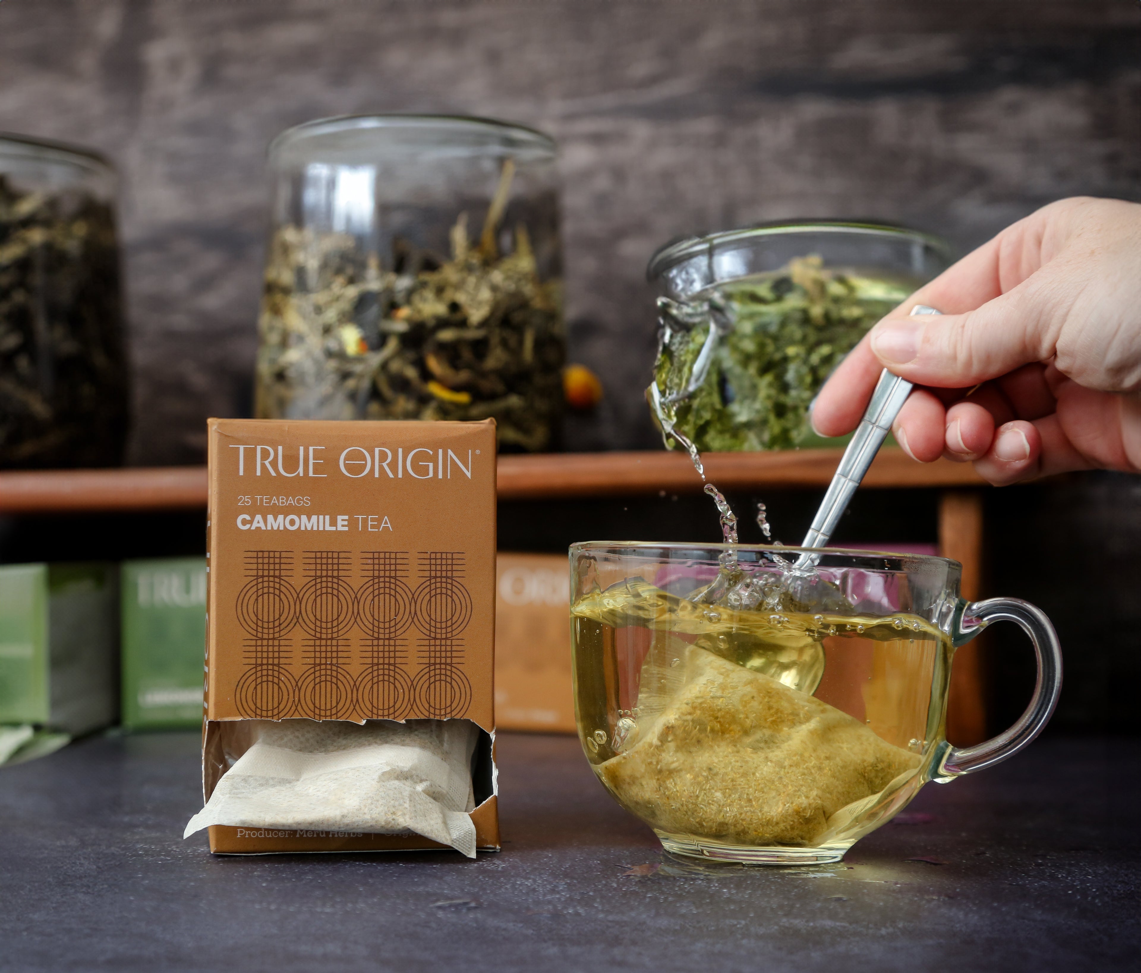A box of True Origin Camomile Tea with 25 tea bags, producers name and origin (Kenya) printed on it.  Next to someone stirring camomile tea in a clear.