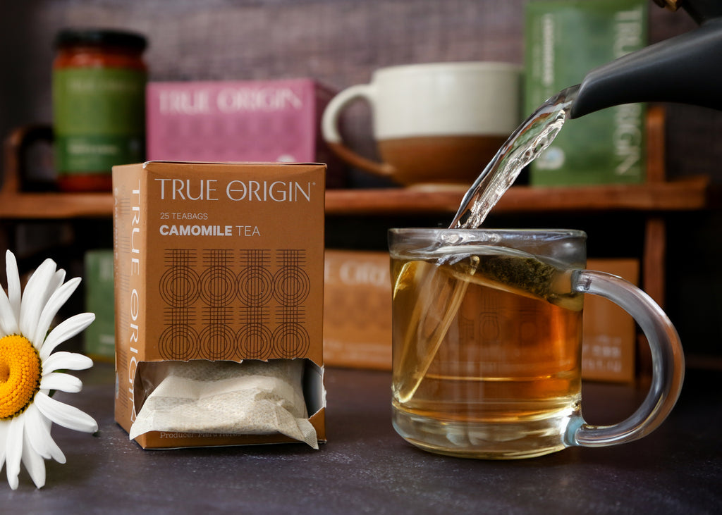 A box of True Origin Camomile Tea with 25 tea bags, producers name and origin (Kenya) printed on it, next to some water being poured from a teapot into a clear mug of camomile tea.