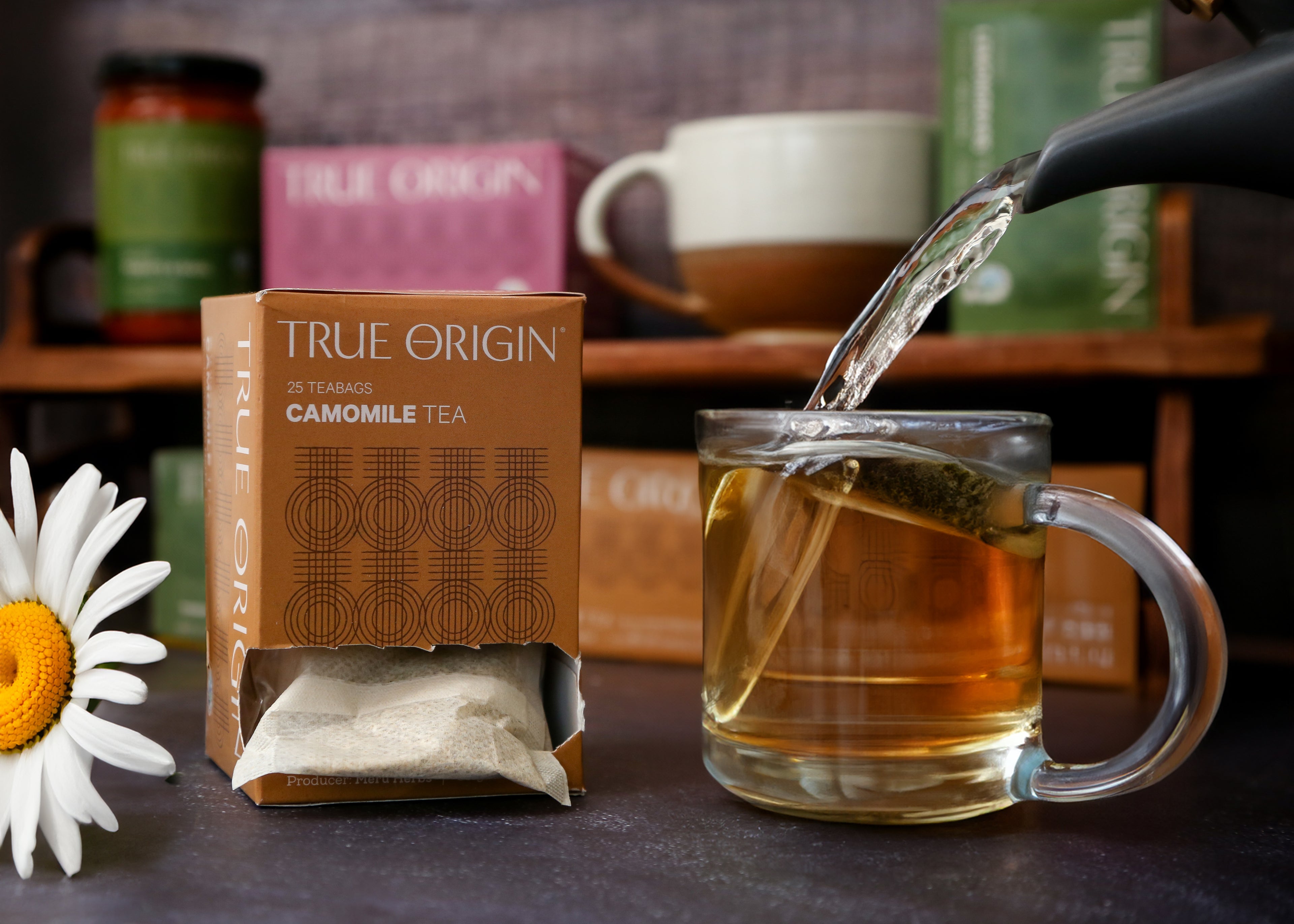 A box of True Origin Camomile Tea with 25 tea bags, producers name and origin (Kenya) printed on it, next to some water being poured from a teapot into a clear mug of camomile tea.