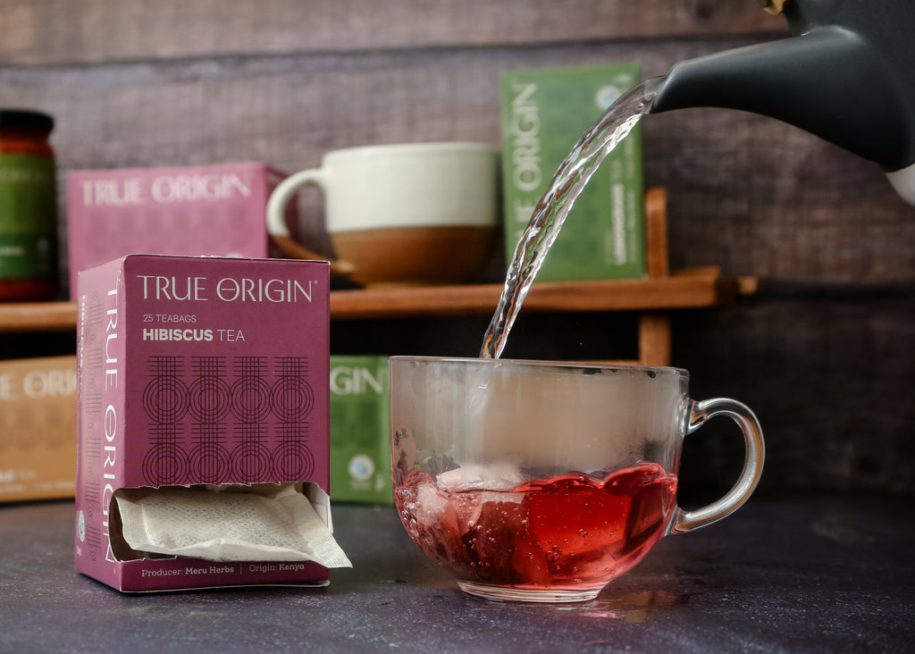 An open box of True Origin brand hibiscus tea bags, with a purple and magenta carton and some boiling water being poured from a teapot into a clear cup with a hibiscus teabag in it.