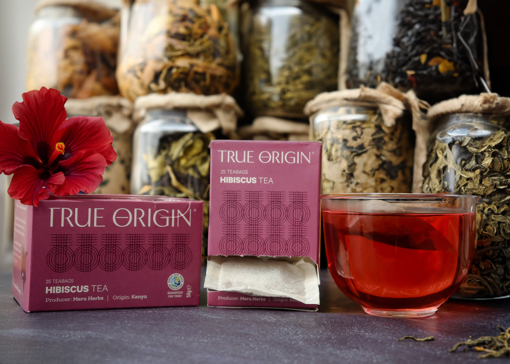An open box of True Origin brand hibiscus tea bags, with a purple and magenta carton next to an unopened box and a cup of hibiscus tea in a clear cup.