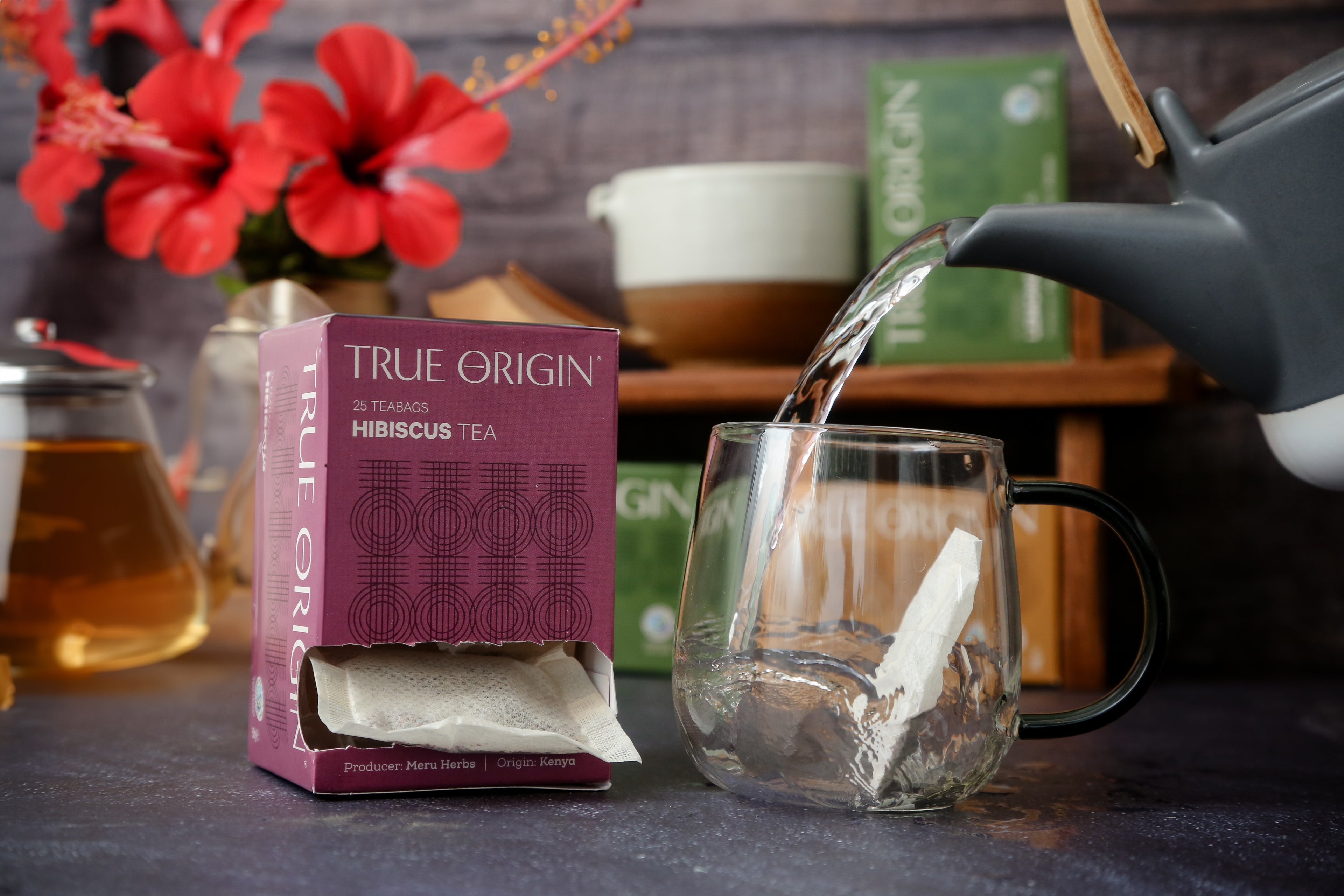An open box of True Origin brand hibiscus tea bags, with a purple and magenta carton next to water being poured from a teapot into a clear cup with a hibiscus teabag in it.