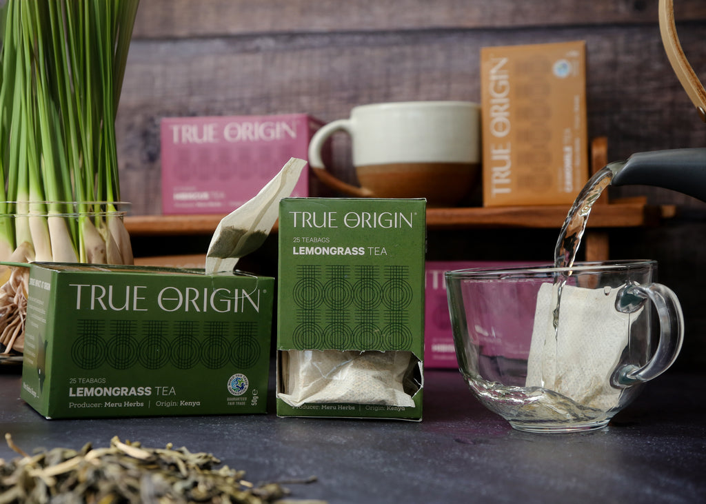 An opened box of True Origin Lemongrass Tea beside an unopened box and some water being poured from a teapot into a clear mug containing a lemongrass teabag.