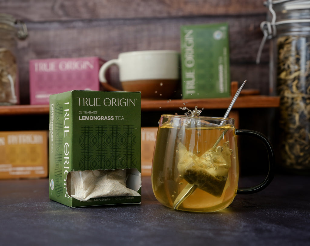 An opened box of True Origin Lemongrass Tea with 25 tea bags, produced by Meru Herbs, origin Kenya.  It is sitting next to a clear mug of lemongrass tea.