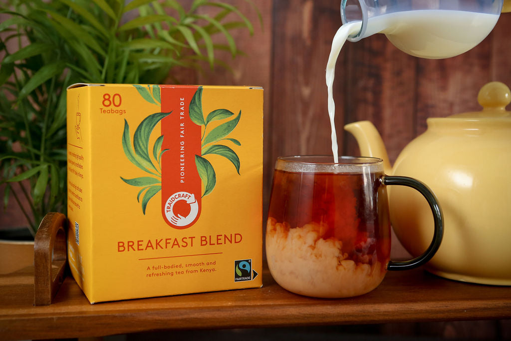 Fairtrade Traidcraft Tea bag packaging labelled 'Breakfast Blend' with a cup of tea being poured into it, set against a wooden surface with a plant in the background.