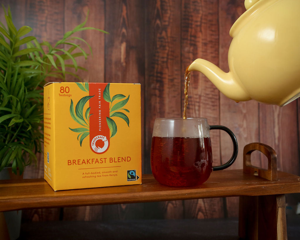 Yellow teapot pouring Fairtrade Traidcraft tea into a glass mug next to a box of 'Breakfast Blend' tea on a wooden surface.