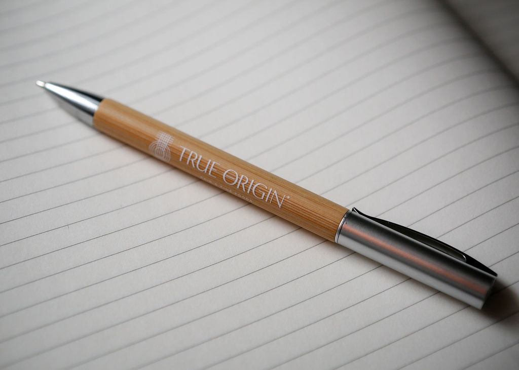 A bamboo and silver pen with a white True Origin logo and symbol lying on a lined notebook.