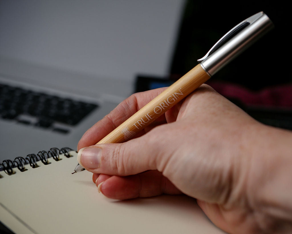 A hand writing with a bamboo and silver pen with a white True Origin logo and symbol.