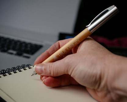 A hand writing with a bamboo and silver pen with a white True Origin logo and symbol.