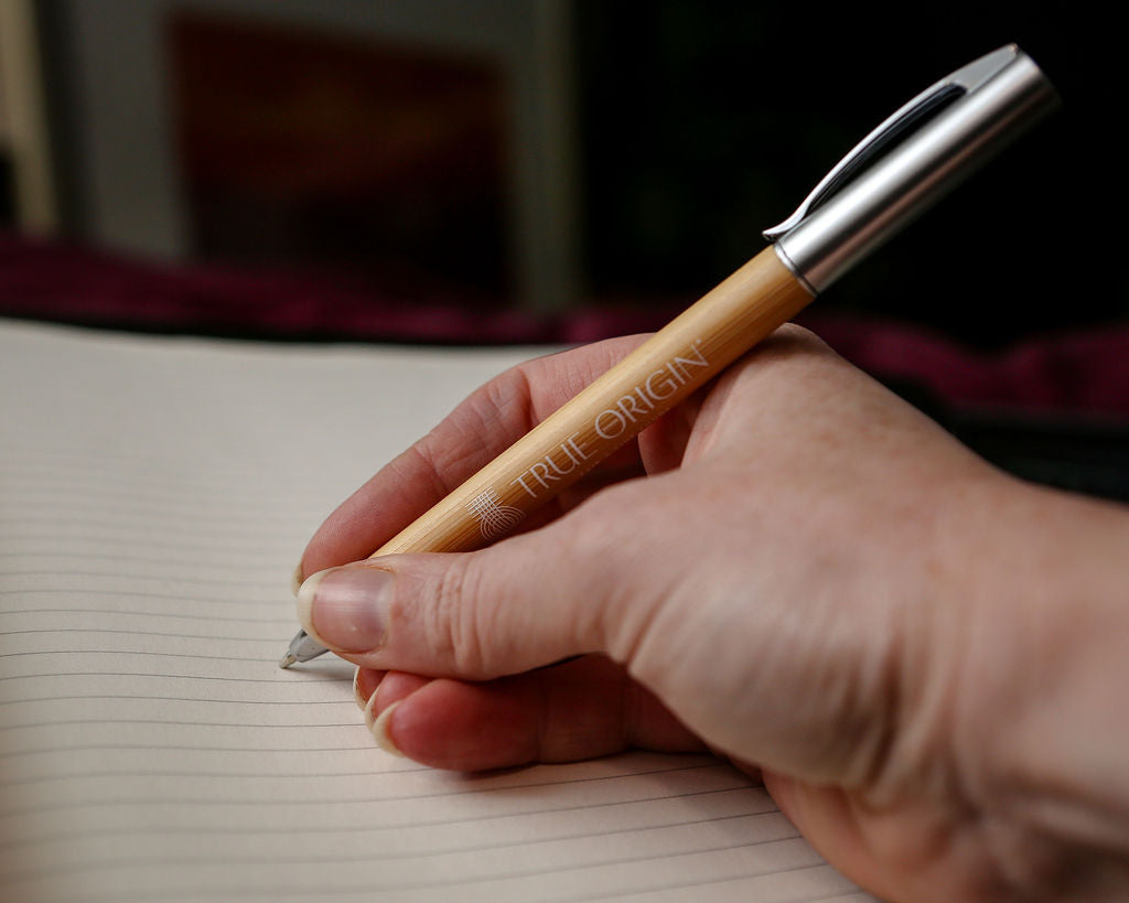 A hand writing with a bamboo and silver pen with a white True Origin logo and symbol.