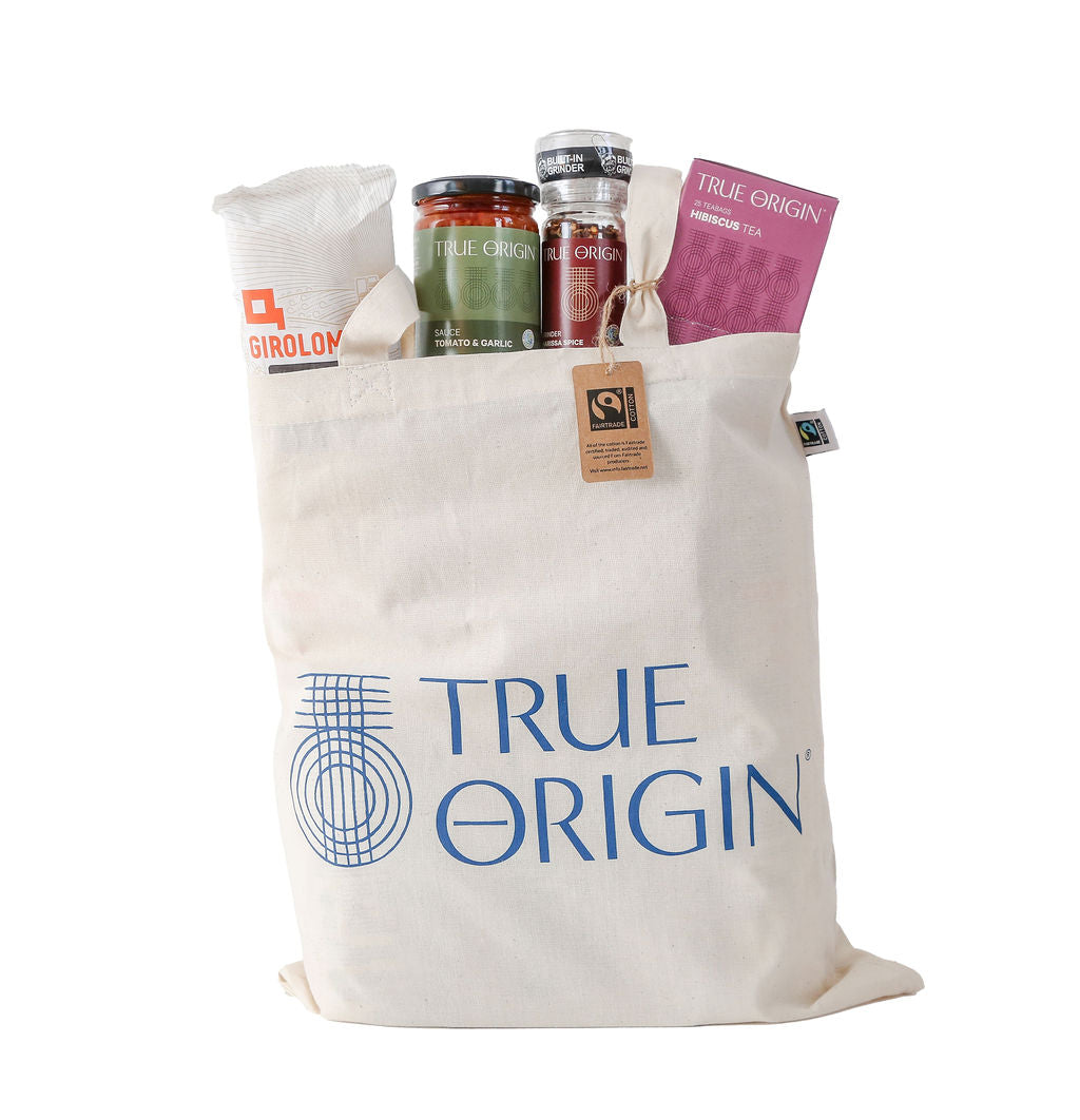 A cream Fairtrade cotton tote bag with the blue stacked True Origin logo with some True Origin products sticking out the top.