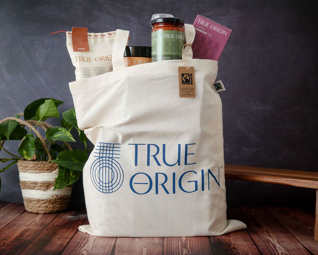 A cream Fairtrade cotton tote bag with the blue stacked True Origin logo with some True Origin products sticking out the top.