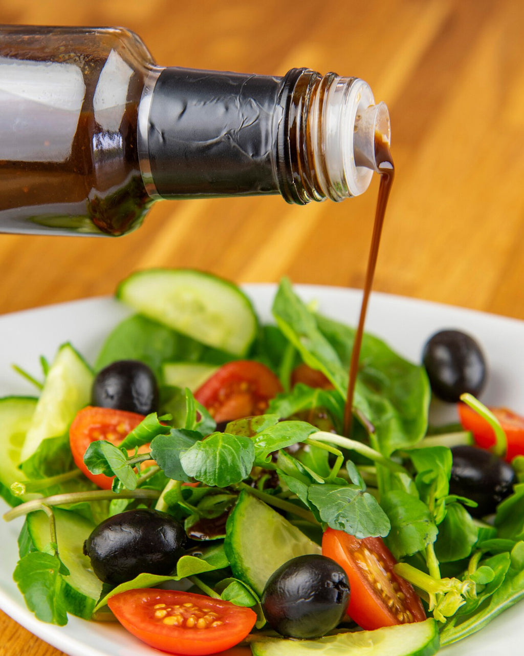 A bottle of Sundried Fig Balsamic Reduction is shown pouring a dark, rich liquid over a salad of sun-ripened figs, walnuts, and cheese on a wooden surface.