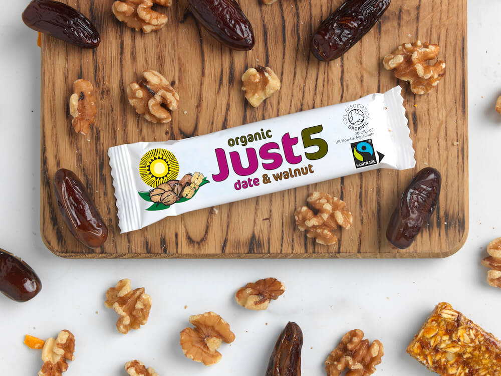 A Fairtrade and organic Just5 date and walnut snack bar on a chopping board surrounded by fresh dates and walnuts.