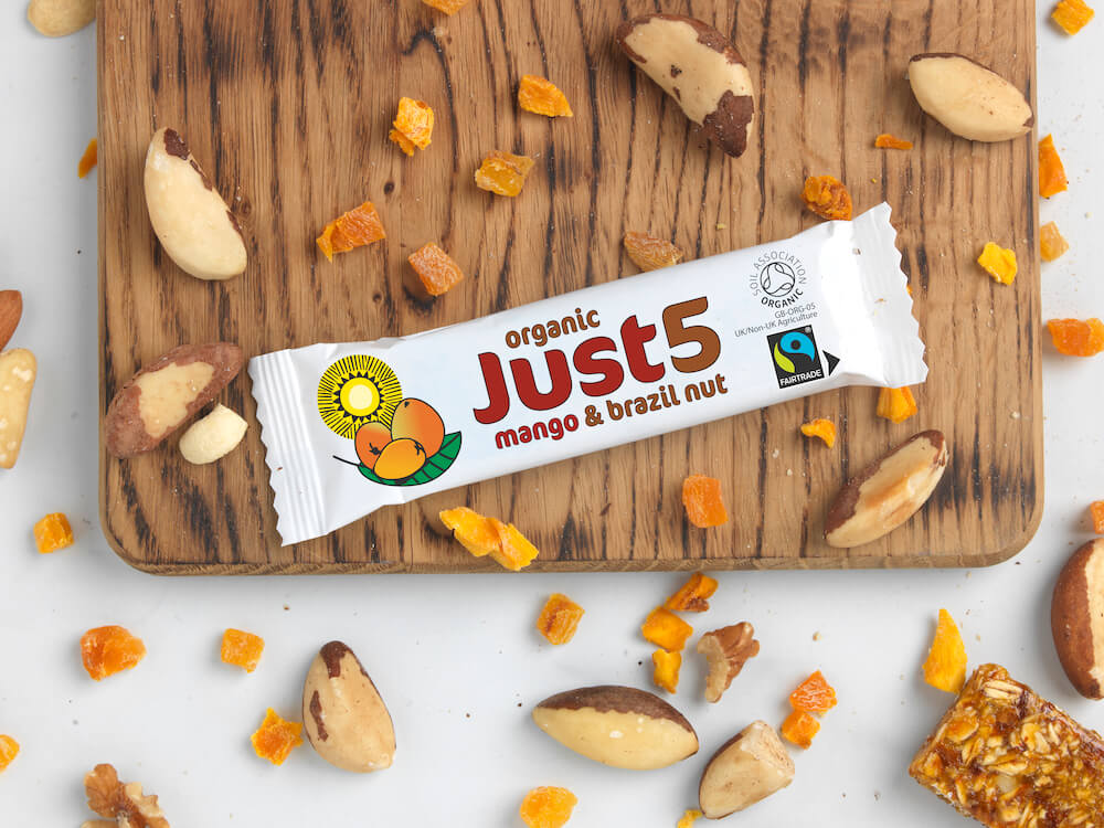 A Fairtrade and organic Just5 mango and brazil nut snack bar on a chopping board surrounded by dried mango pieces and raw brazil nuts.